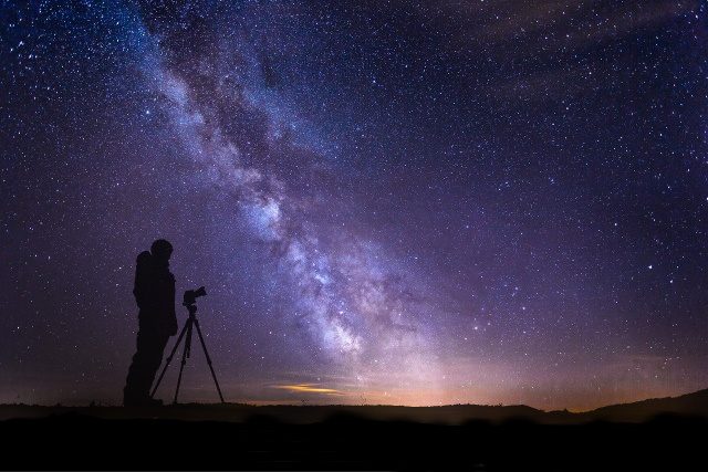 Image of  a scenery showing the Milky Way galaxy arm and a silhouette of a human and a camera on tripod.